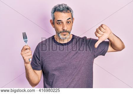 Handsome middle age man with grey hair holding one silver fork with angry face, negative sign showing dislike with thumbs down, rejection concept 