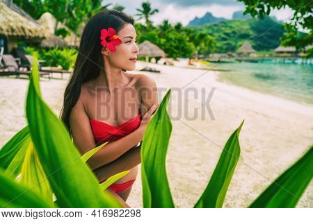 Natural exotic beauty woman in red swimsuit looking serene on tropical beach. Asian bikini model wearing hibicus flower for body care pampering.