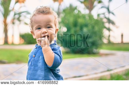 Sad little boy putting fingers on mouth touching gums because toothache at the park on a sunny day. Beautiful blonde hair male toddler in pain for new baby teeth outdoors