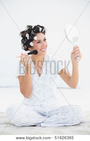 Cheerful brunette in hair rollers holding hand mirror and applying makeup at home in bedroom