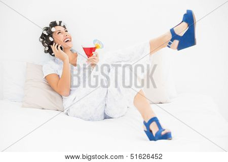 Brunette in hair rollers and wedge shoes holding a cocktail making a call on bed in bedroom at home