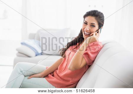 Laughing brunette sitting on her sofa on the phone looking at camera at home in the living room