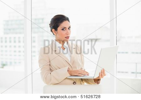 Frowning businesswoman standing behind her chair holding laptop in her office