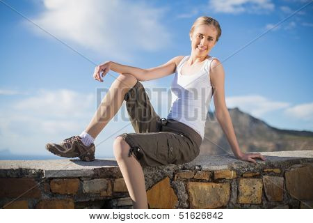 Smiling woman sitting on wall looking at camera against mountain and blue sky