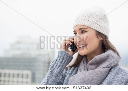 Happy brunette with winter clothes on having a call outdoors on a cold grey day