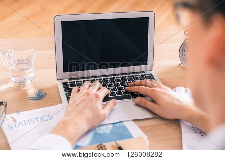 Rearview of male hands using blank black laptop on office desk with business report glass of water and alarm clock. Mock up