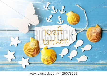 Flat Lay View Of Label With German Text Kraft Tanken Means Relax. Sunny Summer Greeting Card. Butterfly, Shells And Fishes On Blue Wooden Background