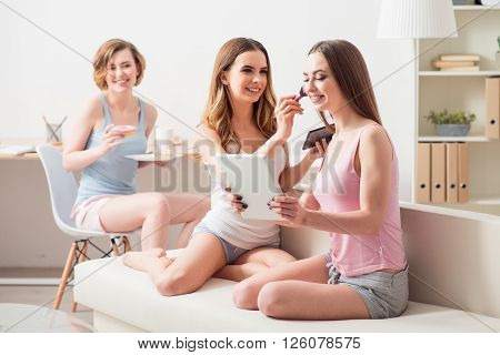 Positive atmosphere. Cheerful delighted beautiful girls sitting on the couch and  doing make up while their friend resting nearby