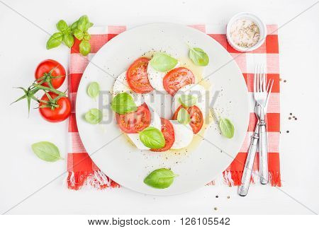 CLassic Italian Caprese salad with tomatoes, mozzarella di Buffala and fresh basil on red and white checkered napkin over white wooden background. Top view