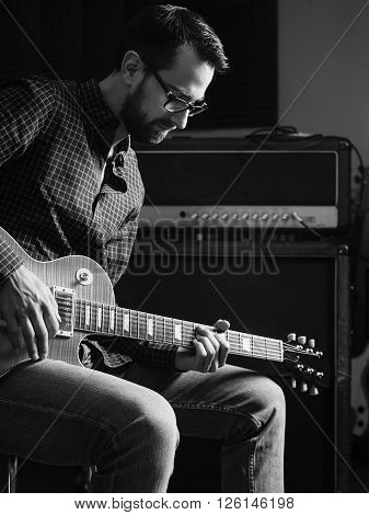 Photo of a man sitting playing his electric guitar in front of a large amplifier.