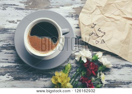 Cup of coffee, flowers and piece of crumpled paper with note I LOVE YOU on wooden table, top view