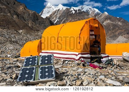 Solar Battery on Rocky Glacier Moraine for Electricity Supply of Sport Mountain Expedition in Wilderness Country with Camping Tents on Background
