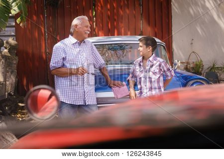 Family and Generation gap. Old grandpa spending time with his grandson. The senior man talks vintage car with the boy and lean against the hood of an automobile from the 60s
