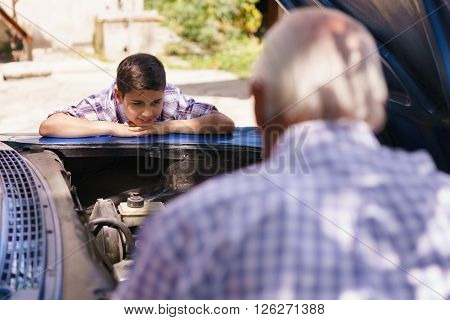 Family and Generation gap. Old grandpa spending time with his grandson. The senior man shows the engine of a vintage car from the 60s to the preteen child. They boy smiles happy.