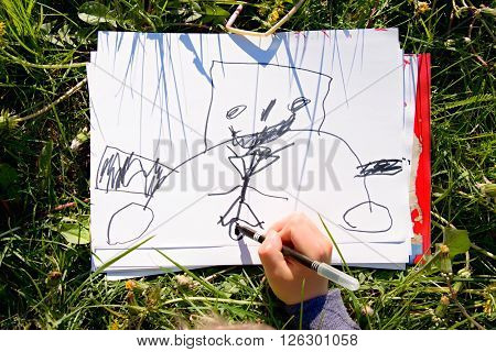 Close Up View Of Child's Hand Drawing Colorful Ornaments On Paper. Notebook In Fresh Green Grass Of
