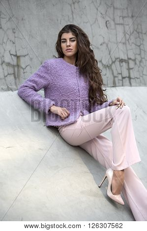 Cute girl with beautiful hair sits on the concrete wall and looks into the camera. She wears violet jacket, pink pants and pink shoes. She holds right hand on her hip, left hand is on the knee of her bent right leg. Outdoors. Vertical.
** Note: Soft Focu
