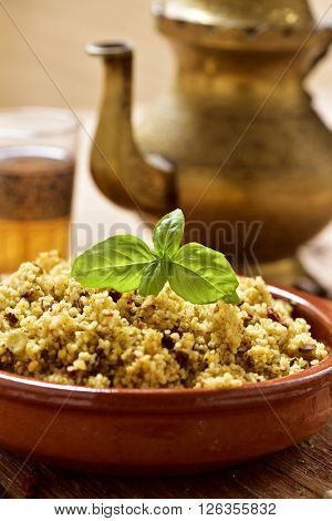 closeup of an earthenware bowl with couscous with vegetables, some ornamented glasses with tea and a golden teapot on a rustic wooden table