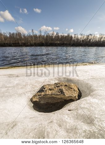 Stone in Ice by River with trees and a patrly cloudy sky
