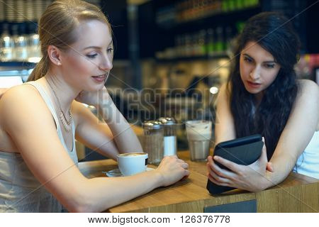 Two young women looking at a digital tablet as they sit enjoying coffee together in a bistro or coffeebar