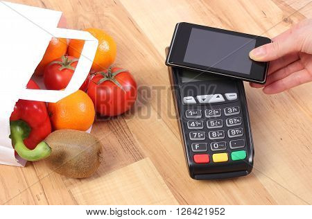 Using credit card reader payment terminal with mobile phone with NFC technology and fresh fruits and vegetables in paper shopping bag cashless paying for shopping