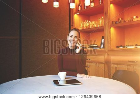Young attractive female is having pleasant mobile phone conversation during rest in contemporary coffee shop modern businesswoman talking on cell telephone during work break in lof office interior