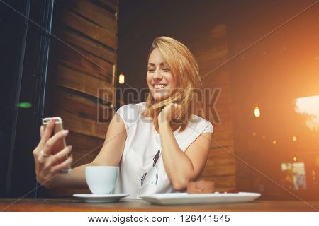 Young cheerful woman posing while photographing herself on smart phone camera for a chat with her friends attractive smiling hipster girl making self portrait on cell telephone while sitting in cafe