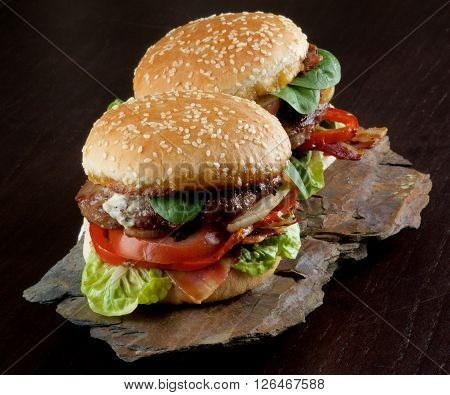 Two Tasty Hamburgers with Beef Bacon Lettuce Tomatoes Basil Roasted Onion and Juicy Sauce on Sesame Buns on Stone Board closeup on Dark Wooden background