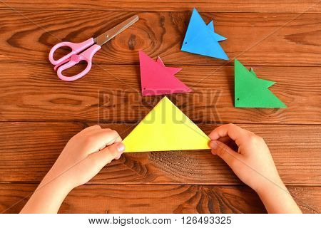 Origami colorful fish, scissors. Child holds paper sheet in his hands and making origami fish. Brown wooden background