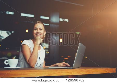 Young female with cute smile sitting with portable net-book in modern coffee shop interior during recreation time charming happy woman student using laptop computer to prepare for the course work
