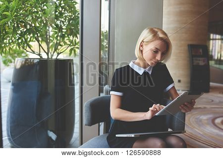 Young elegant female skilled managing director is reading e-mail on digital tablet positive feedback from the client about her work while is sitting in luxury restaurant interior during break at job