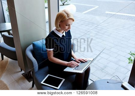 Young female is making on-line purchase via laptop computer while is sitting in modern co-working space. Businesswoman is checking e-mail in internet via portable net-book during coffee break in cafe