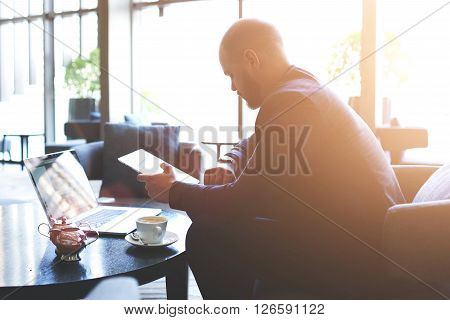 Young man skilled CEO is checking time while is waiting with touch pad and net-book international partners. Male restaurant owner with portable digital tablet in hand is looking on his wrist watch