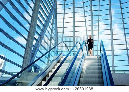 Female skilled CEO is dialing a number on mobile phone while is standing near escalator in interior with contemporary design. Woman is using cell telephone while is moving on staircase in office