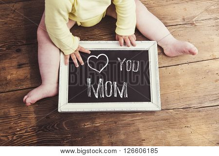 Mothers day composition. Unrecognizable baby in yellow onesie holding a chalk sign in picture frame. Studio shot on wooden background.