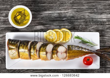 Slices of Smoked fish Mackerel or Scomber on a white rectangular dish with cherry tomato sliced lemon and rosemary on an old wooden table small bowl with olive oil studio lights top view
