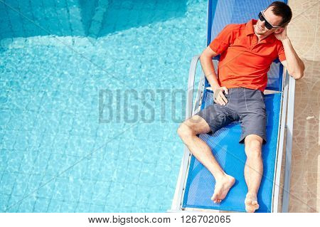 young man relaxing on the chaise-longue near the pool. man wearing in red polo shirt and sunglass