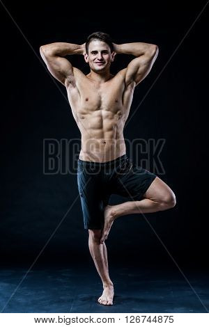 Muscular man balancing on the one leg feeling relaxed on dark background