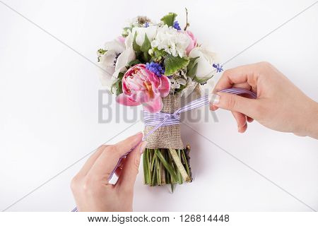Two hands are making a bow from a violet ribbon in beautiful Easter bouquet from pink tulips violet grape hyacinths white anemones violet veronica and white buttercups on white background
