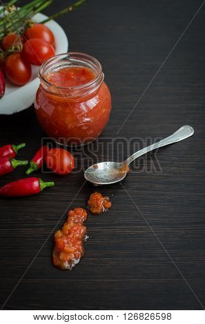 Home made ketchup in a jar. Tomato ketchup sauce with cherry tomatoes and mini red hot chili peppers in a small glass jar with a spoon on dark wooden background. Selective focus. Copy space.