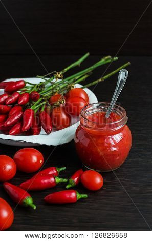 Glass jar of homemade tomato sauce with fresh ingredients on dark wooden background. Natural homemade sauce of tomatoes, peppers and vegetables. Chilli jam. Selective focus.