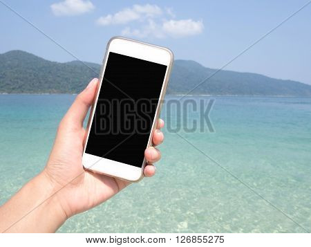 Close up of woman's hand holding smartphone mobile smart phone over blurred beautiful blue sea and clear sky on beach background for working online on vacation. Phone with black screen.
