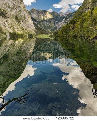 Reflection of blue sky and mountains in calm mountain lake with trees impressive white clouds with view on some stones under water near the shore and a valley in the background