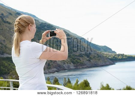 Young beautiful blonde woman doing a picture with your mobile phone. A woman is standing against a background of sea and mountains she is a tourist. Photograph