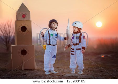 Two adorable children boy brothers playing in park on sunset dressed like astronauts imagining they are flying on the moon