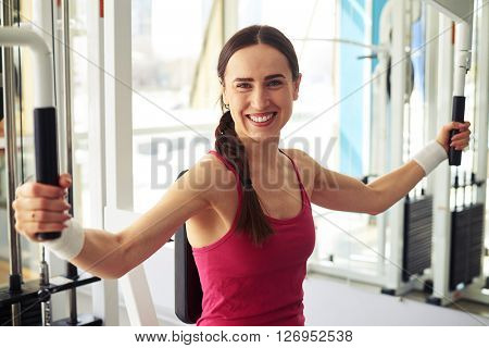 Beautiful Caucasian smiling girl is working out on butterfly machine in gym near window