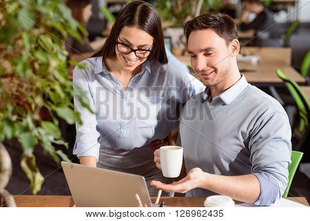 Help me. Cheerful handsom smiling man drinking coffee and sitting at the table while using laptop with his colleague who is standing nearby
