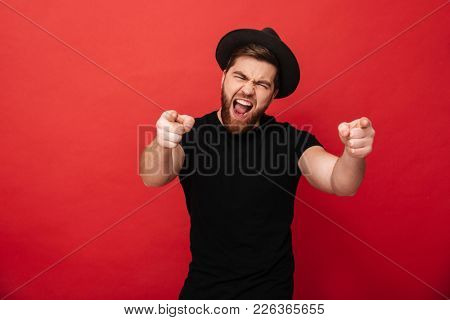 Photo of emotional fancy guy wearing black t-shirt and hat having fun and pointing fingers on camera meaning hey you isolated over red background