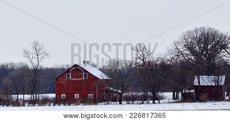 A Valleyside Red Barn On A Farmstead Appears On A Snowy Day.