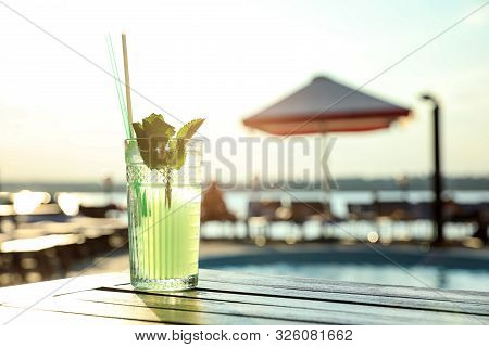 Glass Of Fresh Summer Cocktail On Wooden Table Near Swimming Pool Outdoors At Sunset. Space For Text