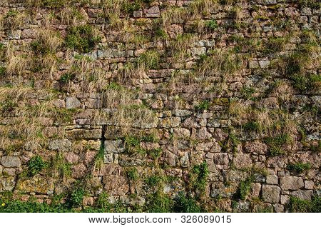Fragment Of An Old Fortress Wall. The Wall Is Made Of Stone Blocks And Is Overgrown With Grass. Semu